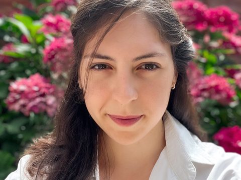 A head and shoulders photograph of Magalie, long hair in a white shirt, standing in front of beautiful flowers