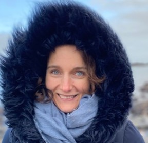 Emily Thomas in a furry hood standing on a beach with sea in the background